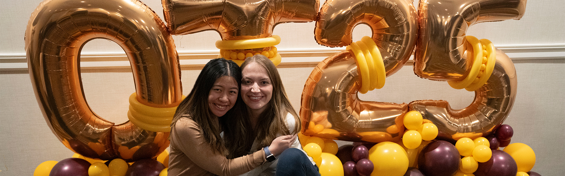 Two women embracing in front of balloons that spell out "OT 25"