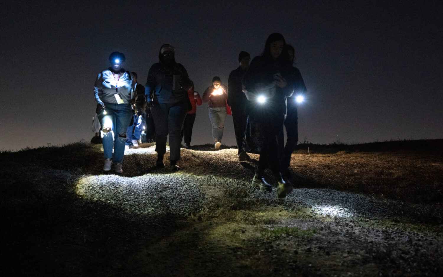 A group of students with flashlights traverse the grounds at night.