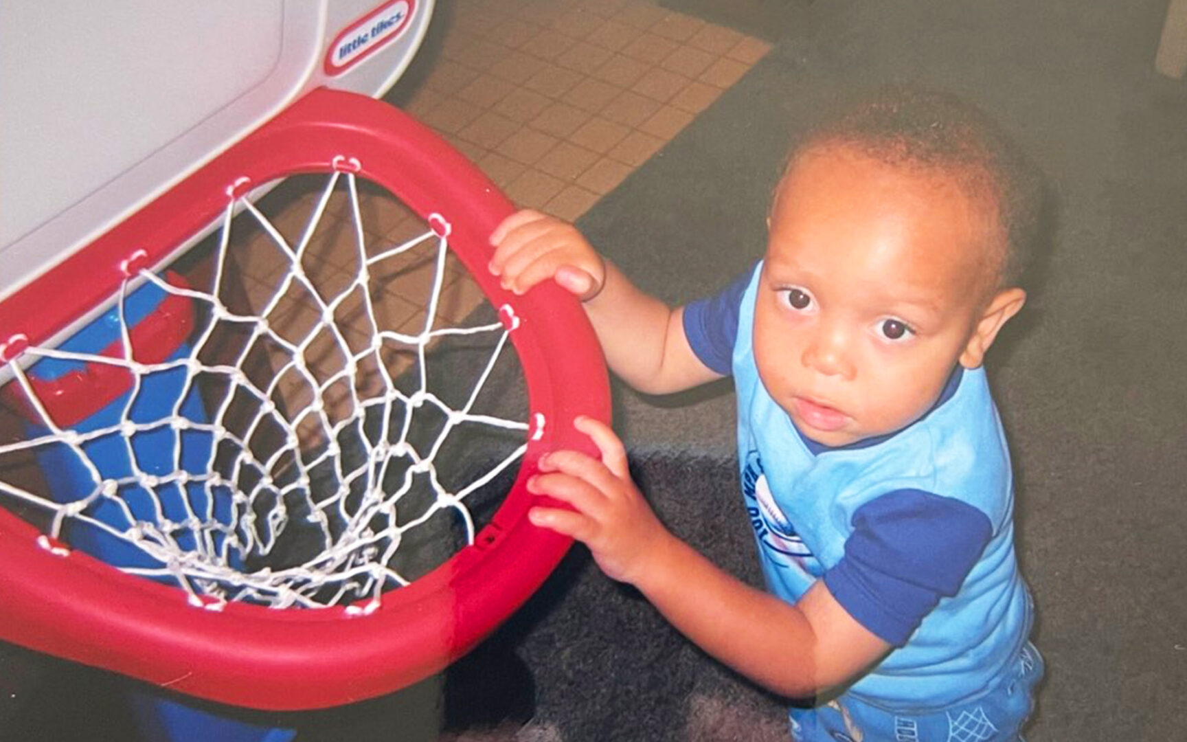 Jeremy as a toddler, grasping a child-sized basketball hoop.